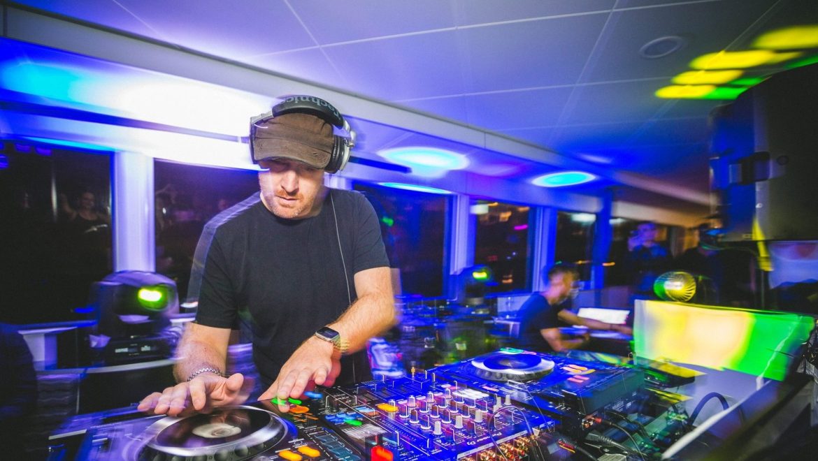 A blurry action shot of a man DJing in a room with lights and CD-J equipment.