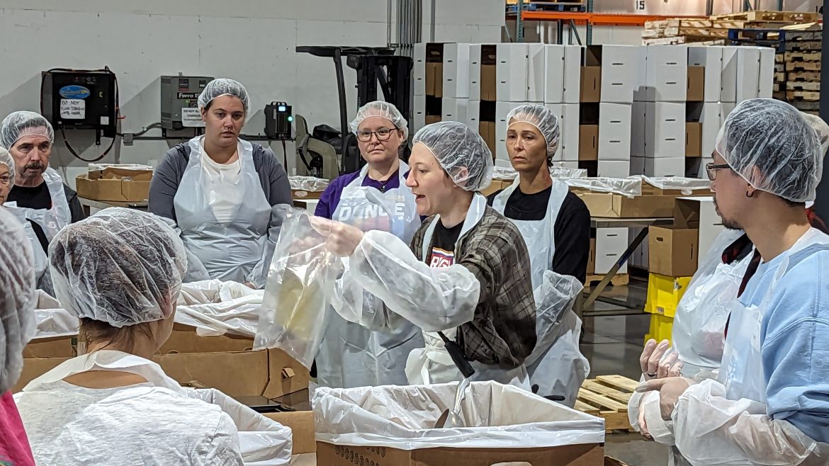 A woman explaining to a group of people in PPE how to pack food.